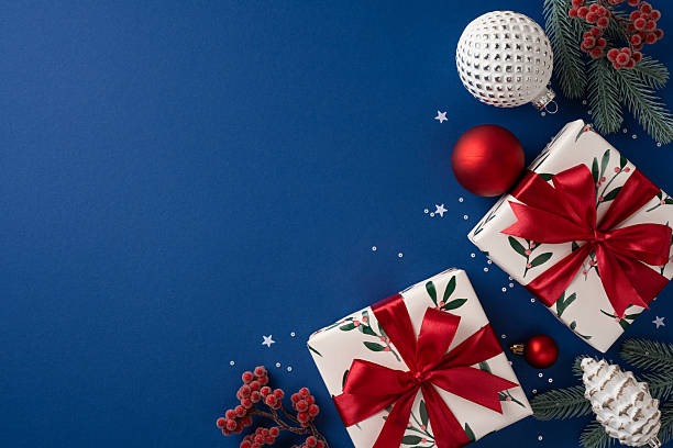istock Decorative gifts with leafy patterns and red bows rest beside ornaments and pine sprigs on a deep blue backdrop, conveying holiday joy and seasonal warmth for Christmas and New Year projects 2237400406