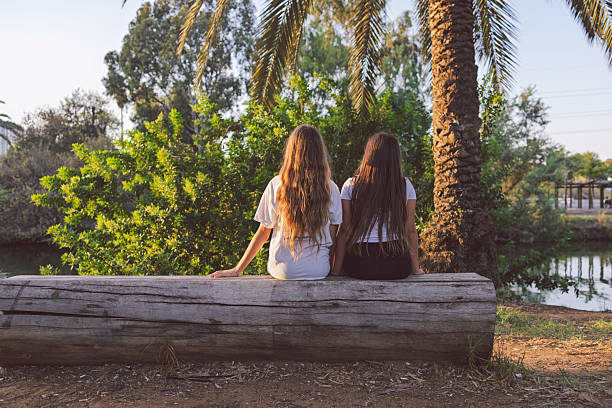 Two girls sitting on a wooden log by a lake in a park, viewed from behind