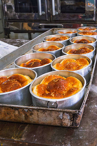 Rows of warm, golden brown bread rolls in round metal pans resting on a rustic bakery table after baking.