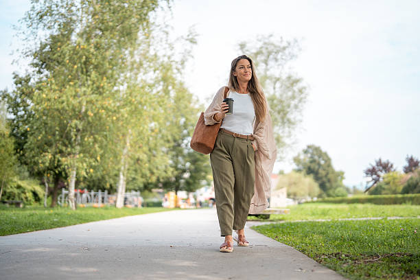 Mid adult Caucasian woman walking on a sunny day in a park, holding a coffee and carrying a bag. She is wearing casual attire and enjoying the peaceful, green surroundings featuring lush trees and open pathways.
