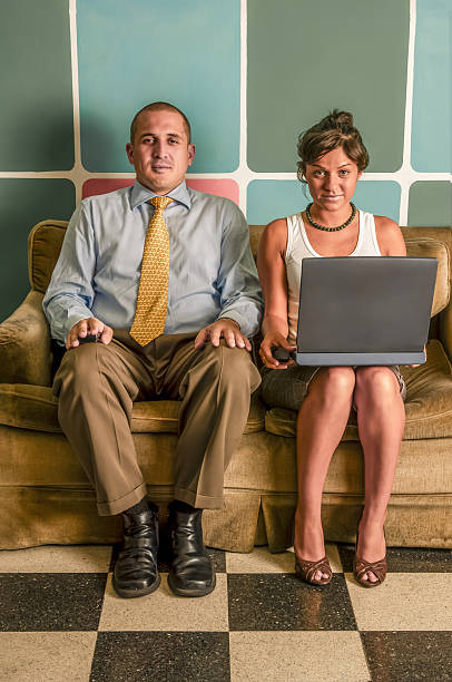 Two young business professionals sitting on a vintage sofa in an office, with the woman balancing a laptop on her lap, engaging in teamwork and communication indoors. Concept of a retro old fahioned workspace.