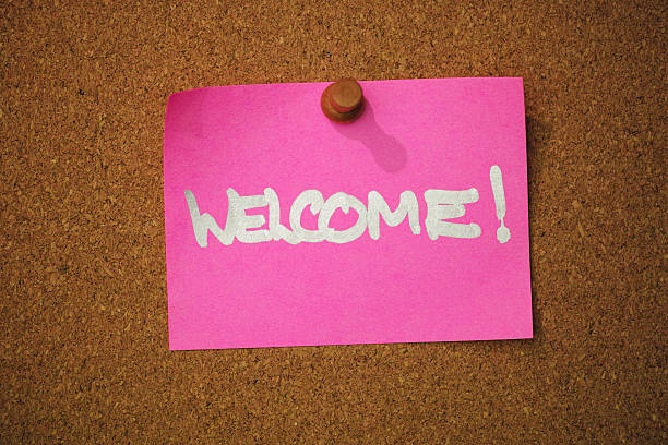 The word 'WELCOME !' written in a silver marker pen on pink note paper, pinned by a wooden pin to a cork message board. Conceptual greeting message relating to welcome, etc.