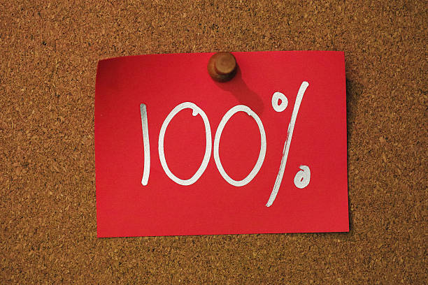 The number '100%' written in a silver marker pen on red note paper, pinned by a wooden pin to a cork message board. Conceptual message relating to one hundred percent, quality, the whole amount, purity, etc.