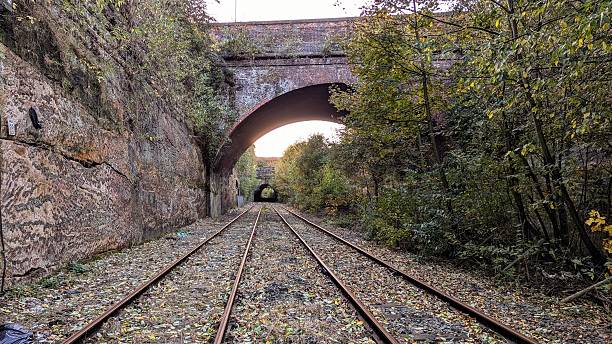 istock Abandoned railway line with parallel straight long train tracks towards horizon. Overgrown mountain cliffs pass along side by side rails and sleepers littered with autumn leaves, footbridge gravel. Sunset visible in distance beneath arched railroad bridge 2237440403