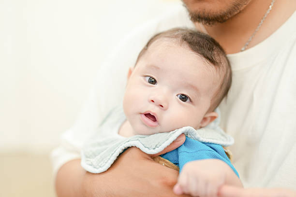 A baby is being held by a man in a blue shirt. The baby has a blue bib on and is wearing a blue shirt. The baby is looking at the camera with a surprised expression