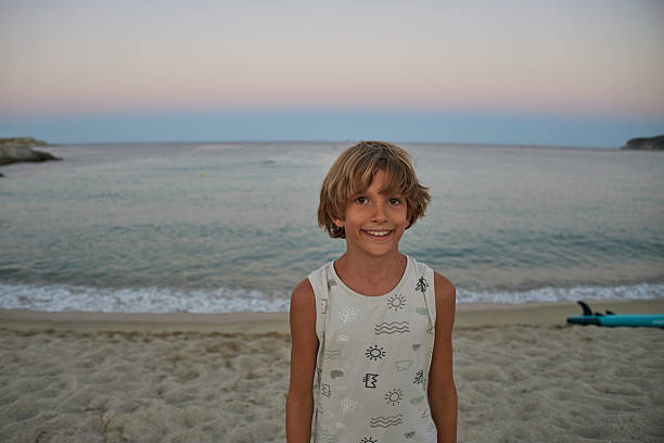 Young boy standing on a sandy beach during twilight, looking at camera and smiling