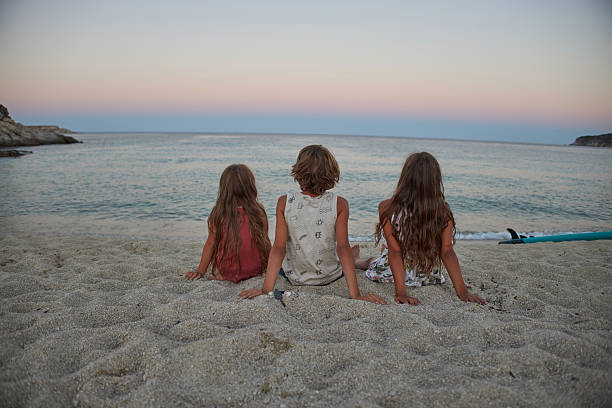 Three children relaxing on a sandy beach, enjoying the quiet evening sky