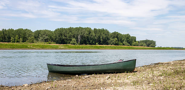 istock Calm Canoe Resting on Sandy Riverbank Under Open Sky with Green Trees in Background 2237447616