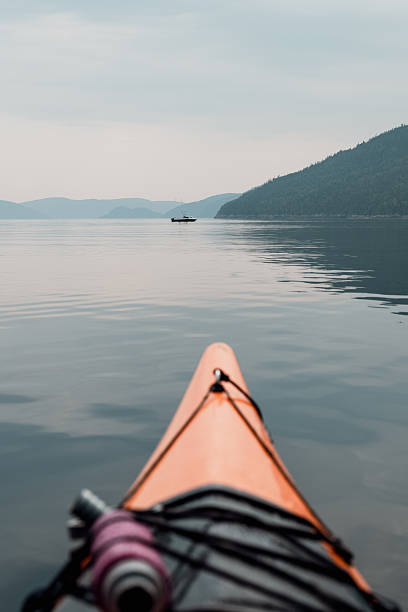 First-person perspective from an orange kayak gliding over calm waters toward a distant boat, with a mountain coastline and soft sky creating a tranquil, adventurous mood.