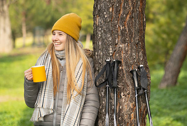 Hiker drinking hot beverage leaning against tree after nordic walking