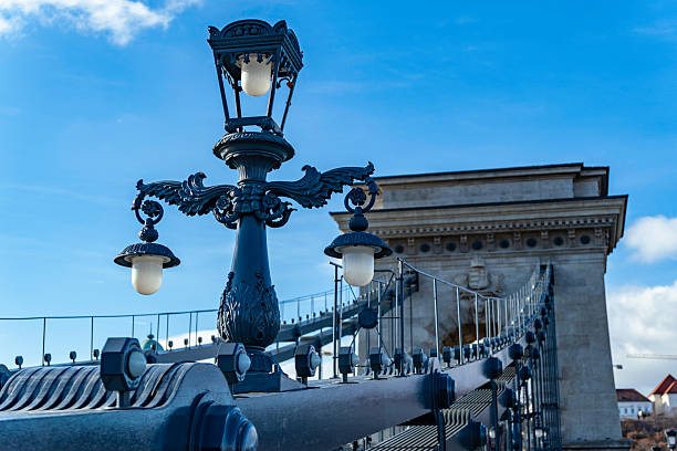 Close-up view of the ornate cast-iron lamp post and suspension cables on the historic Széchenyi Chain Bridge in Budapest, Hungary. The iconic 19th-century landmark, spanning the Danube River, is a symbol of Hungarian architecture and culture, captured under a bright blue sky.