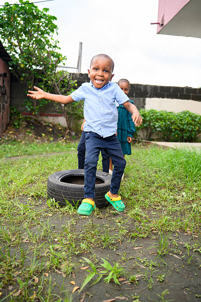 A young boy in a blue shirt and jeans jumps on grassy ground beside a black car tire, smiling joyfully, with other children in the background.