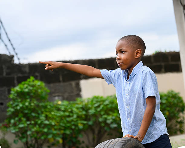 Young boy in a light blue shirt and dark pants points outward with a serious expression while holding a tire in a grassy yard with trees and a wall in the background.