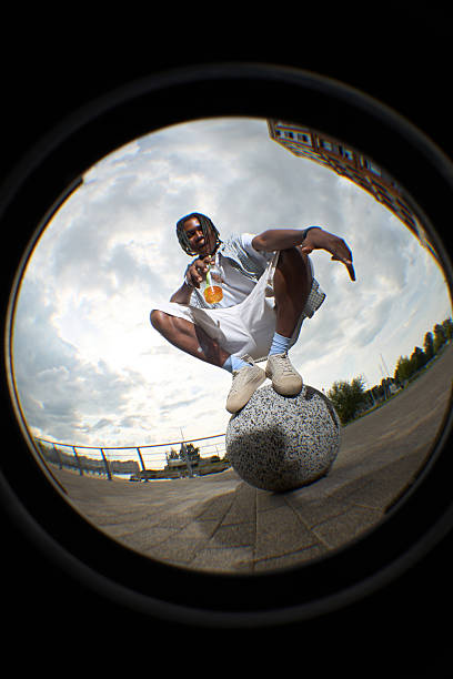 Black young man crouching on large stone sphere outdoors, holding drink in plastic cup, looking confidently at camera, urban background visible through fisheye lens