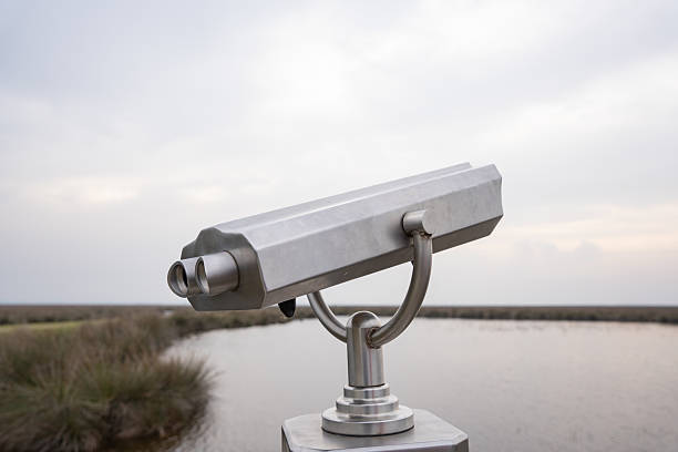 Fixed silver binoculars on a sturdy metal stand are positioned near a small water body, set in a grassy landscape. The overcast sky casts a diffused light, enhancing the serene outdoor scene. The binoculars likely offer visitors a closer view of the surrounding area, emphasizing their function as part of a tourist or nature observation point. The remote and natural setting suggests a location that values wildlife observation or landscape viewing.