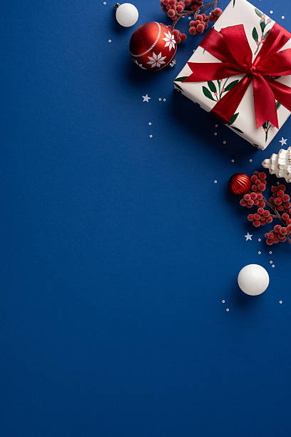 istock A vertical festive scene featuring a wrapped gift with a red ribbon, red ornament, white balls, and berry sprigs on a blue backdrop. Perfect for Christmas, New Year, greeting cards 2237467261