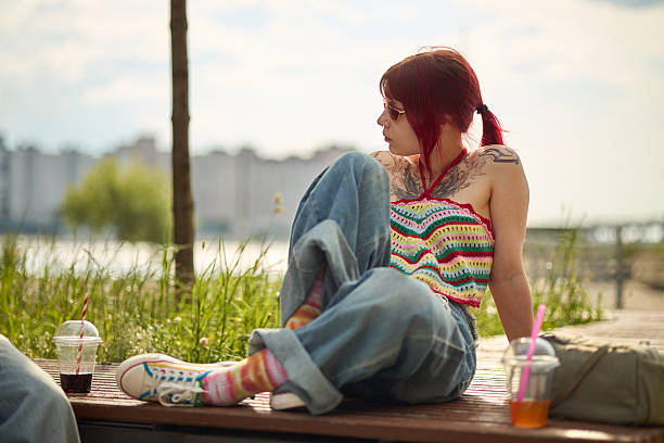 Young Caucasian woman with red hair and visible tattoos sitting on bench outdoors near river, looking away from camera, relaxing with two takeaway drinks on sunny day