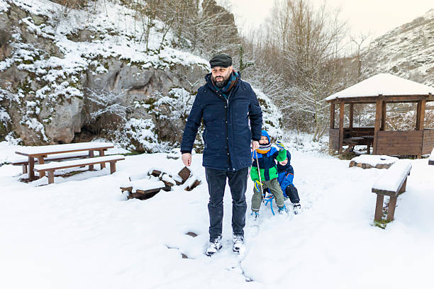 Happy father spending quality time with his children in a snowy outdoor setting, pulling them on a sled through a scenic winter landscape with picnic benches and a wooden gazebo in the background. Family enjoying fun seasonal activities together in nature.