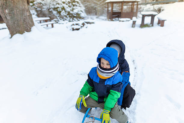 Two cheerful kids bundled up in warm winter clothes sitting together on a sled, ready for a fun ride in a snow-covered park. Outdoor winter adventure and childhood joy on a cold, sunny day.
