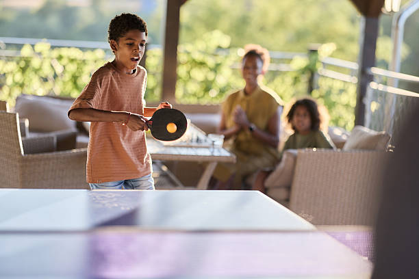 African American boy playing table tennis against someone while mother and girl are in the background.