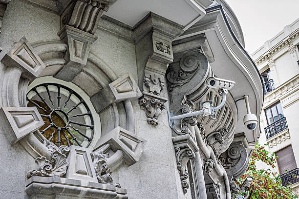 Multiple modern security cameras are installed on the exterior of a classical sandstone building in downtown Madrid, Spain. The facade features ornate architectural details including Corinthian columns, scrollwork, and a circular stained-glass window, creating a striking contrast between old-world craftsmanship and contemporary surveillance technology. The weathered stone surface adds texture and historical depth to the scene, while the presence of cameras reflects current urban safety measures. Ideal for themes of civic infrastructure, architectural juxtaposition, public security, or city life in European capitals.