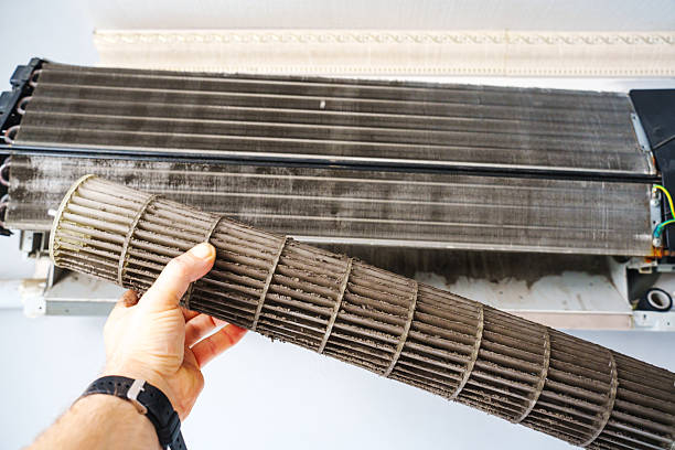 A hand holds a dirty air conditioning filter beside the unit. The filter is covered in dust, emphasizing the need for regular maintenance to ensure good air quality.