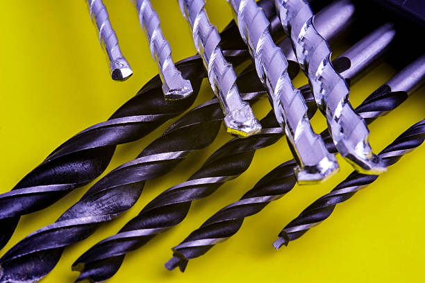 Set of metal drill bits arranged on a bright yellow surface. Both silver and black drill bits with spiral grooves are displayed. Representing essential tools for drilling in wood, metal or masonry.