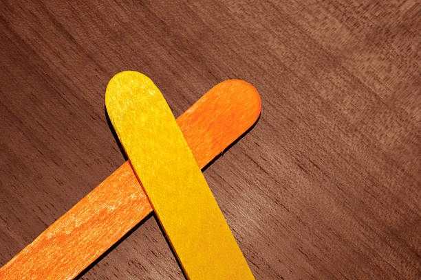 Two colorful wooden craft sticks. One yellow and one orange sticks placed in a crisscross pattern on a polished wooden surface. The warm tones create a simple yet vibrant artistic arrangement.