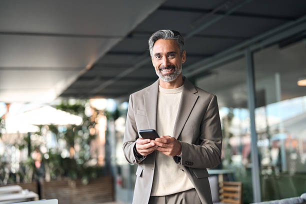 Happy middle aged businessman manager or entrepreneur holding smartphone looking at camera standing outside office. Mature older business man executive using mobile cell phone outdoors. Portrait.
