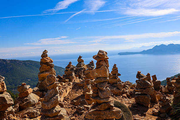 A breathtaking coastal view featuring carefully stacked rock cairns overlooking the Mediterranean Sea. The balanced stone towers stand as symbols of harmony and mindfulness against a backdrop of blue skies, rugged cliffs, and the expansive horizon.
