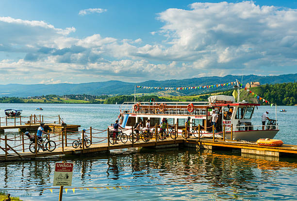Czorsztyn Lake, Poland – August 5, 2025: A tourist boat is docked as cyclists wheel their bikes onto the pier, ready for adventure. The colorful decorations, mountains, and castle in the distance set a lively summer scene.