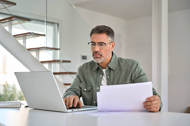 istock Serious mature man looking at paper bill working at home using laptop computer. 2237503414