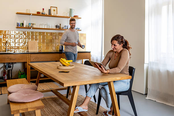 istock Casual Morning At Home Dining Table: Woman Reading Tablet While Man Makes Coffee In Bright Kitchen 2237511322
