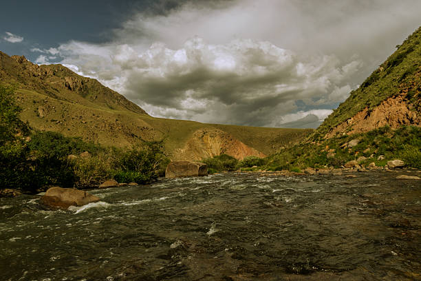 Scenic view of a mountain river flowing among green hills and rocky slopes near Song-Kul Lake, Kyrgyzstan. Summer day with dramatic clouds and untouched wild nature.