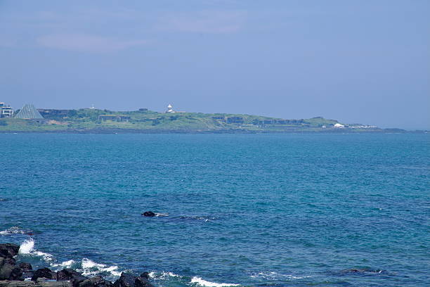 Seopjikoji's white lighthouse stands on the grassy headland beside a distinctive glass pyramid structure, seen across the bay above clear blue water.