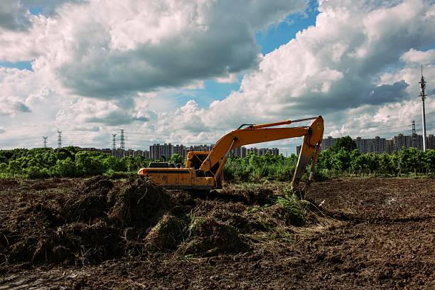This dynamic scene captures a vibrant orange excavator in action, scooping earth on a developing urban site surrounded by lush green trees and scattered soil mounds, with modern high-rise apartments, power lines, and a partly cloudy blue sky in the background. The contrast between industrial machinery and natural elements highlights themes of city growth and environmental integration, perfect for construction, real estate, infrastructure, or urban development visuals in editorial, advertising, or corporate projects.