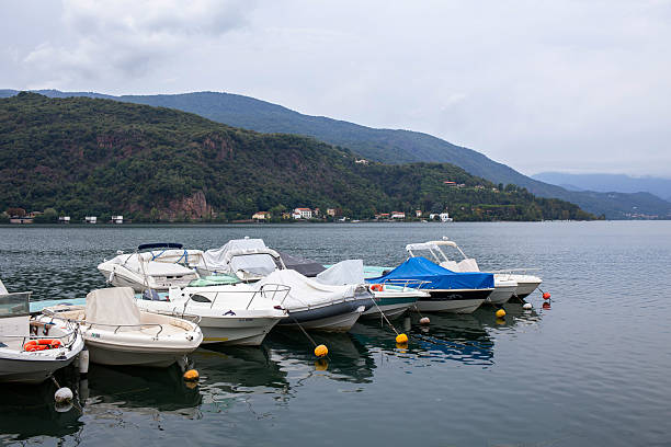 Row of white motorboats moored in a calm lake with a scenic view of green hills and mountains under a cloudy sky.