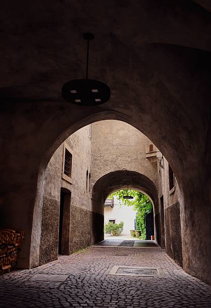 A charming cobblestone alleyway with archways and rustic walls leading to a sunlit courtyard with greenery.