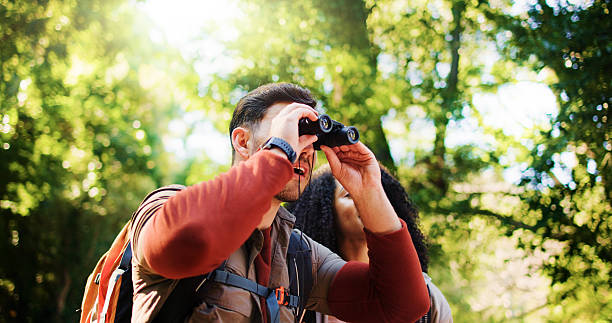 istock Explore, hike and couple in forest with binocular, bird watching or bonding in holiday hobby. Observation, nature and tourist in woods with scope, romantic vacation or animal search in outdoor trip. 2237546941