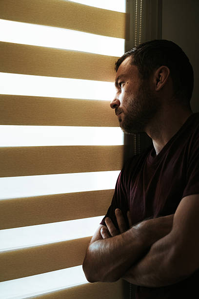 istock Depressed man looking through zebra blinds in dark room at home 2237559378