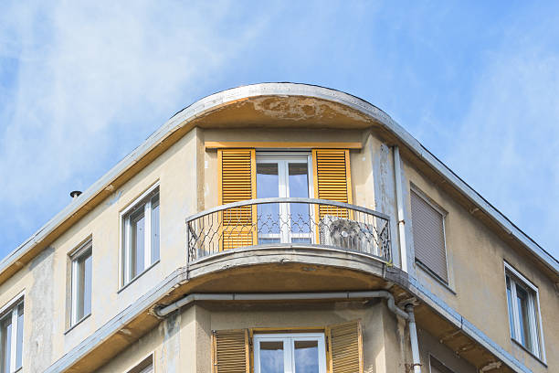 An upward shot showcases the corner of a weathered building featuring a curved balcony with yellow shutters. The architecture is complemented by a partially cloudy sky, creating a contrast between the aged facade and the bright sky.