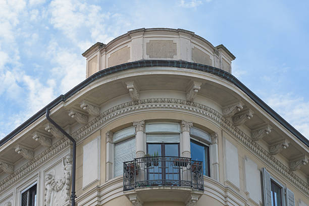 This photograph captures the ornate corner of a historical building featuring intricate architectural details and a wrought iron balcony. The facade showcases architectural styles from 1914, with decorative carvings and a view of a light blue sky with scattered clouds, evoking a sense of timeless elegance and historical significance.