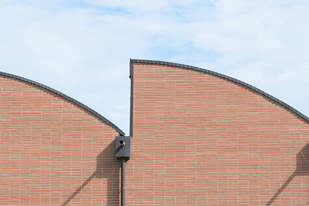 A close-up view showcases a building's facade featuring a sawtooth roof pattern, constructed with red brick. The clear blue sky with wispy clouds creates a clean and modern architectural composition. A downspout is affixed to the building.
