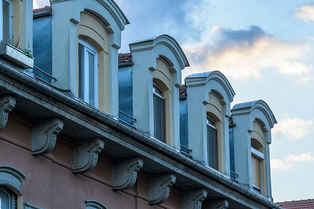 Close-up of a building facade featuring arched dormer windows against a backdrop of a serene sky with soft clouds. The architectural details, including the arched windows and decorative elements, create a classic European aesthetic.