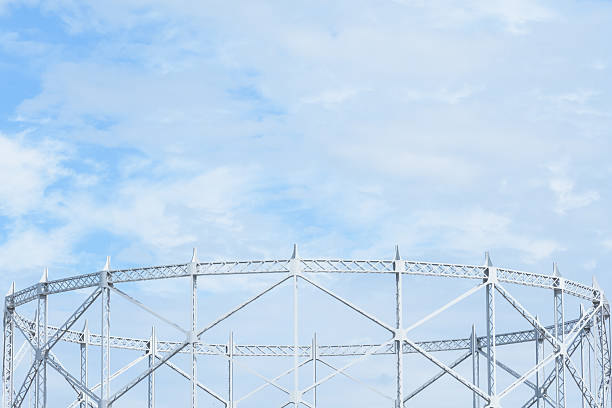An industrial gas holder structure stands prominently against a vibrant blue sky filled with fluffy white clouds. The metal framework, painted in a light color, creates a geometric pattern that contrasts beautifully with the natural sky, offering a unique architectural perspective and sense of space.