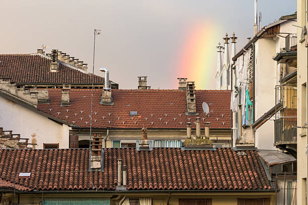 This photograph captures a rainbow arching over a collection of buildings with terracotta tile rooftops, chimneys, and antennas in an Italian city. The image showcases the charm of European architecture with a splash of vibrant color against the muted sky, creating a sense of serenity and wonder.