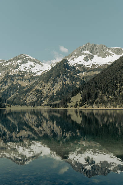 The still waters of Vilsalpsee in Tyrol, Austria, reflect the surrounding mountains in remarkable clarity. Snow remains on the peaks while green meadows and dense pine forests stretch along the shoreline. The lake’s transparency reveals stones beneath its surface, enhancing the layered textures of reflection and depth. A popular hiking destination in the Tannheim Valley, Vilsalpsee is renowned for its untouched alpine scenery and its peaceful atmosphere year-round.
