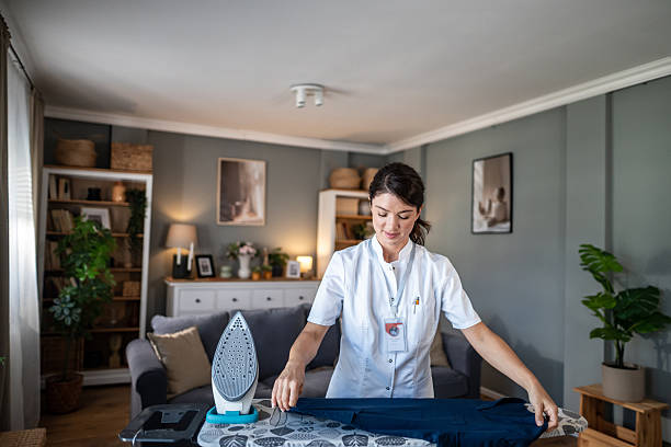 Young adult woman wearing a uniform is performing domestic chores, ironing clothes in a comfortable living room, symbolizing professional home care service and household support
