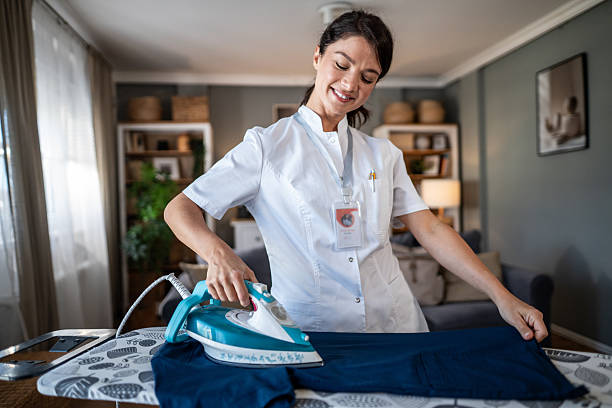 Young adult woman wearing a medical uniform and a smiling expression, ironing her work clothes on an ironing board inside her brightly lit home living room