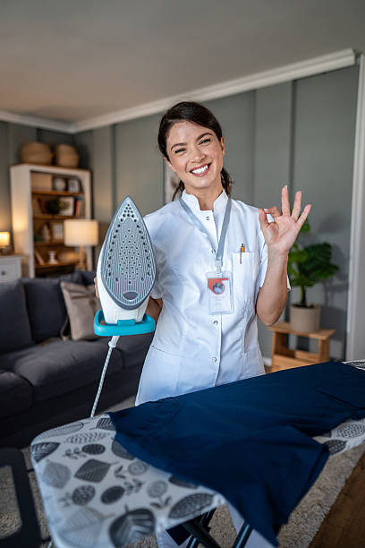 Woman in uniform standing in a living room, holding an iron, making an ok sign, and smiling, perfect for concepts of professional cleaning, domestic services, and home care